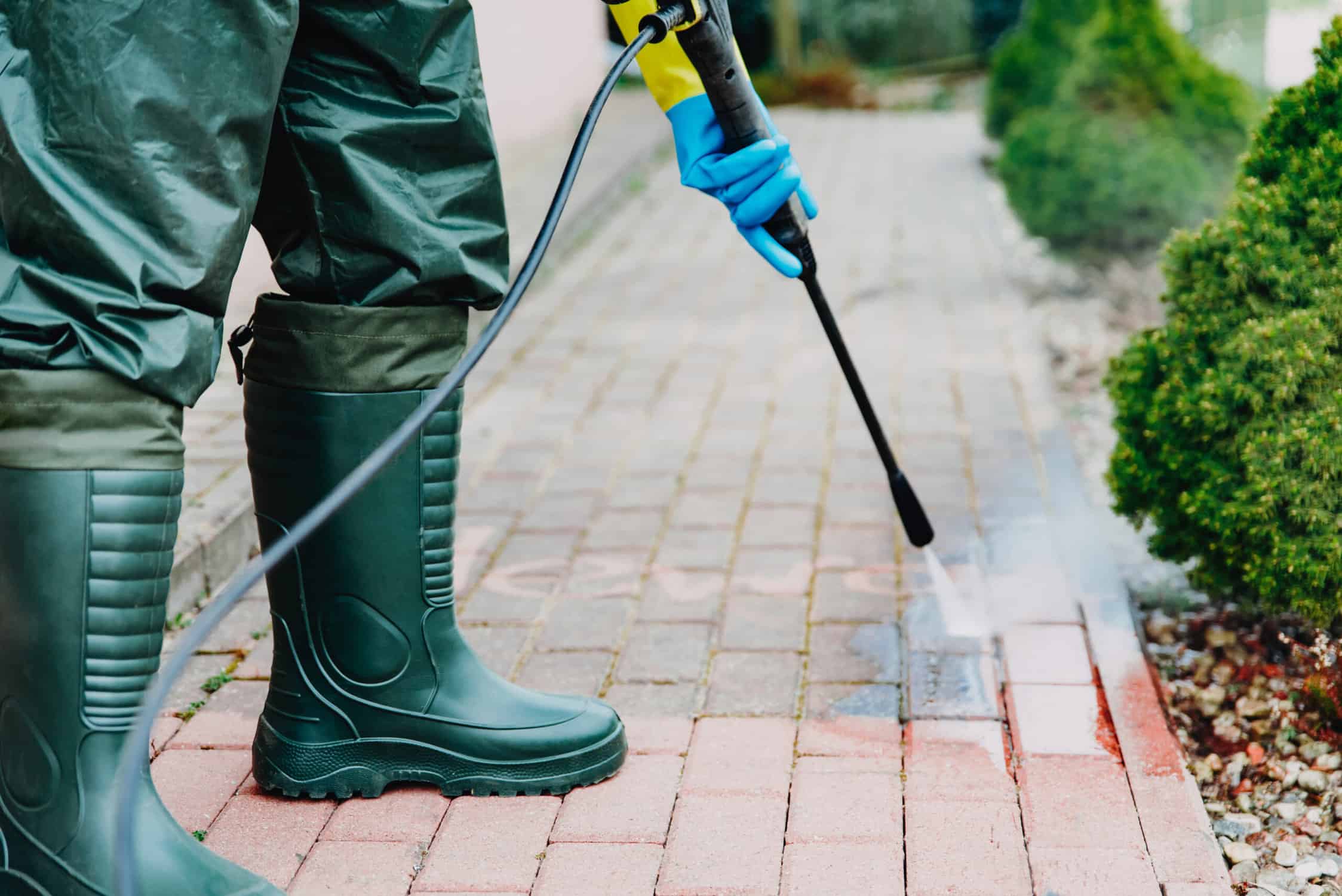 Man cleaning red, conrete pavement block using high pressure water cleaner. High pressure cleaning. Man wearing waders, protective, waterproof trousers and gloves doing spring jobs outdoors.