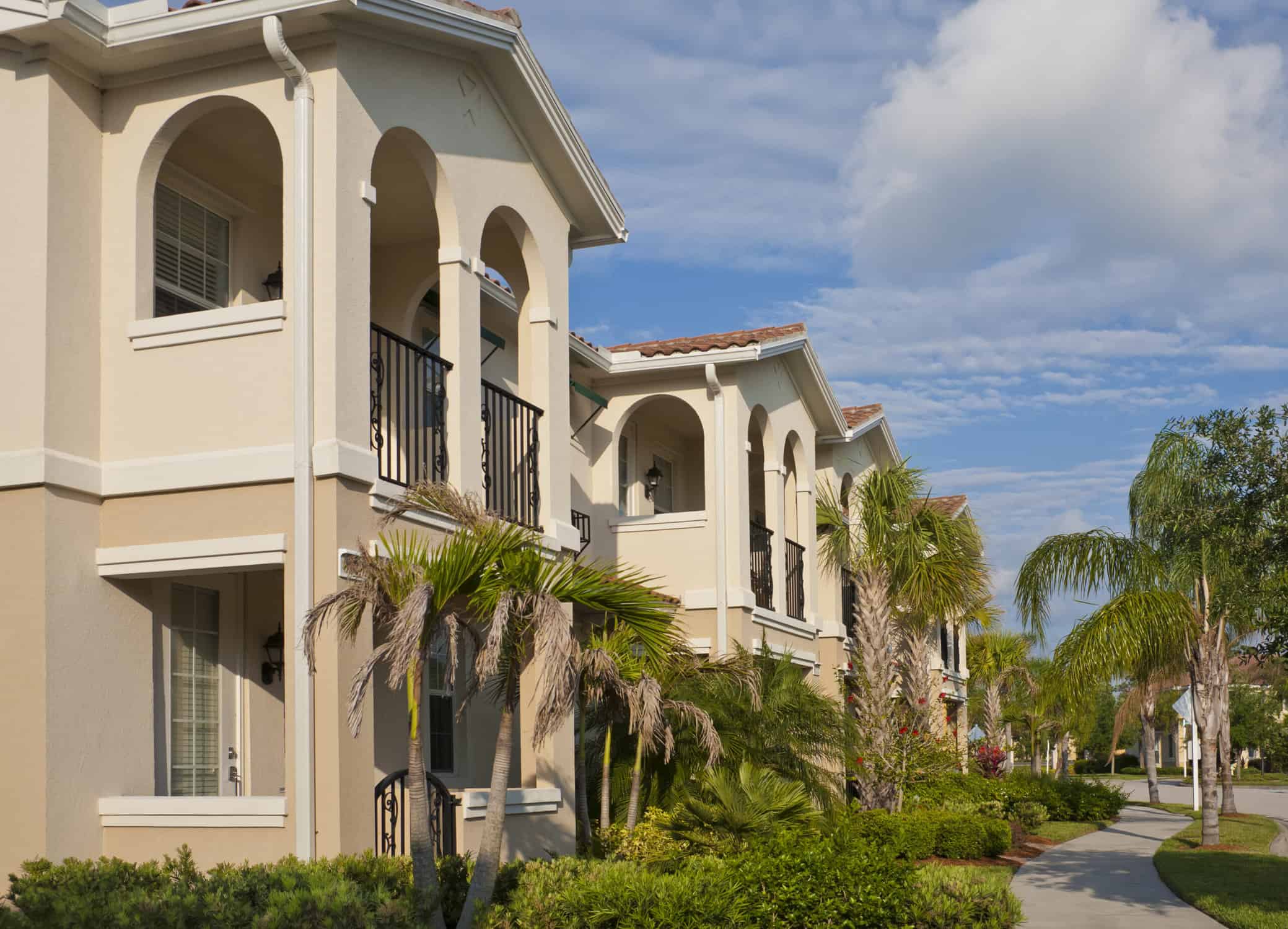 Row of two-story condominiums with sidewalk, palm trees and tropical foliage.