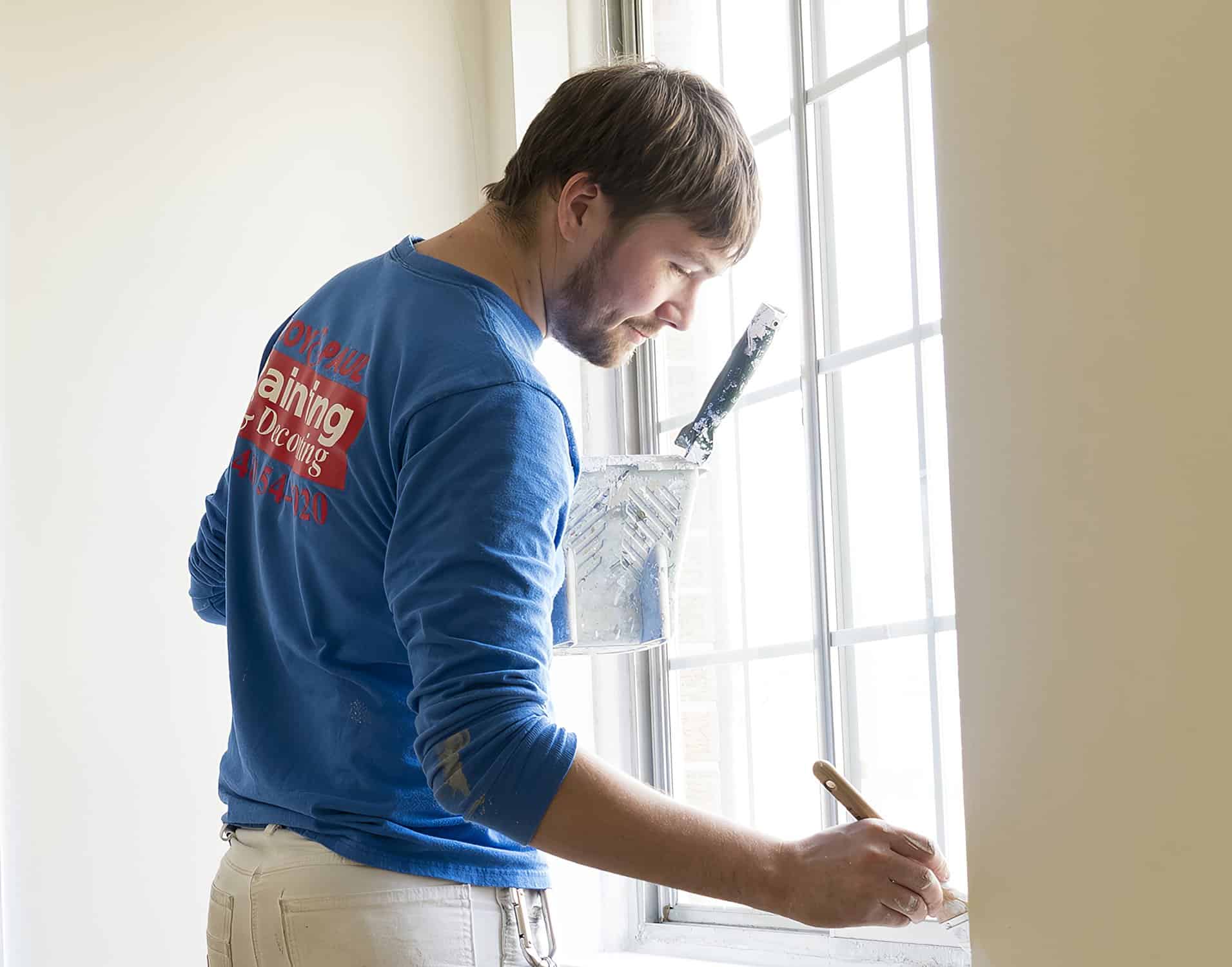 Painter in blue shirt applying cream finish to recessed panel cabinet door frame in Royal Oak