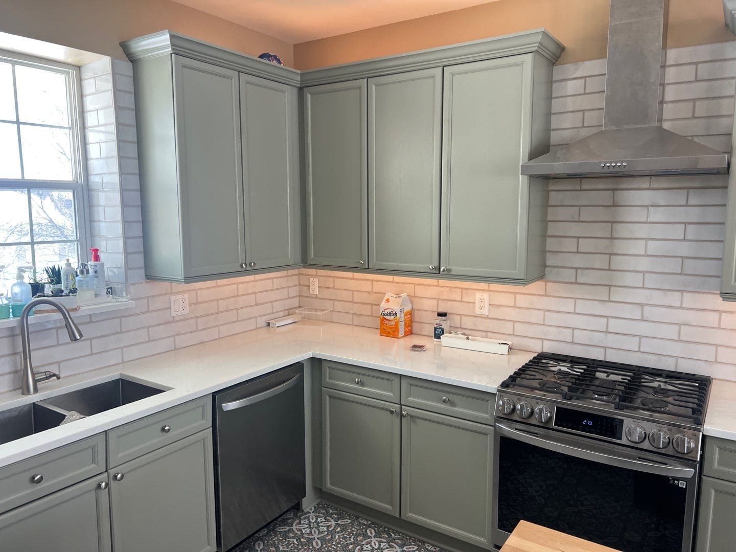 A modern kitchen with freshly painted sage green cabinets, white countertops, subway tile backsplash, and stainless steel appliances.