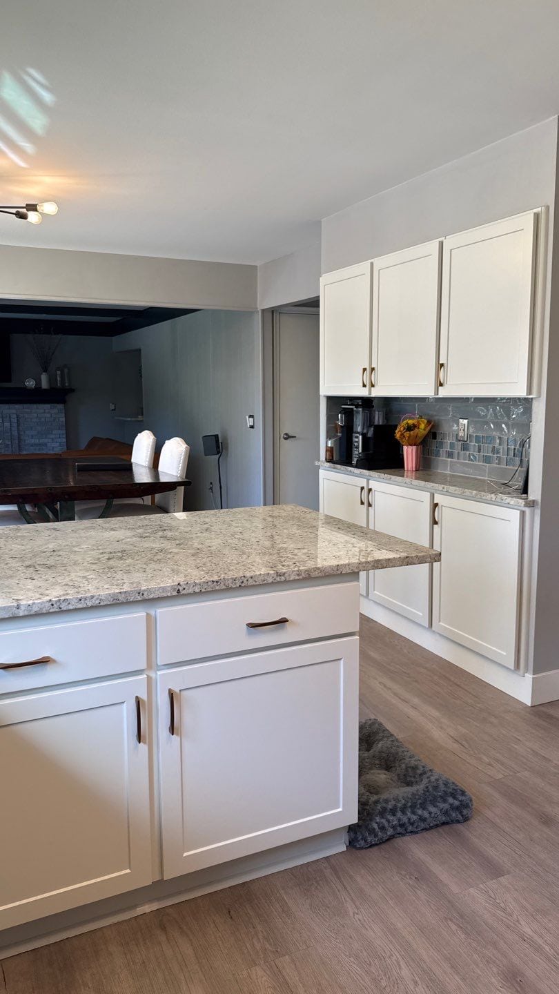 White Painted Kitchen Cabinets Royal Oak White-painted kitchen cabinets with recessed panel doors, bronze hardware, speckled granite countertop, and dark subway tile